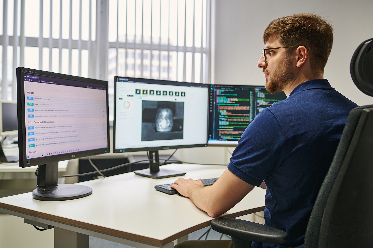 Entwickler am Schreibtisch vor drei Bildschirmen mit API-Dokumentation, optischer Inspektion und Code.