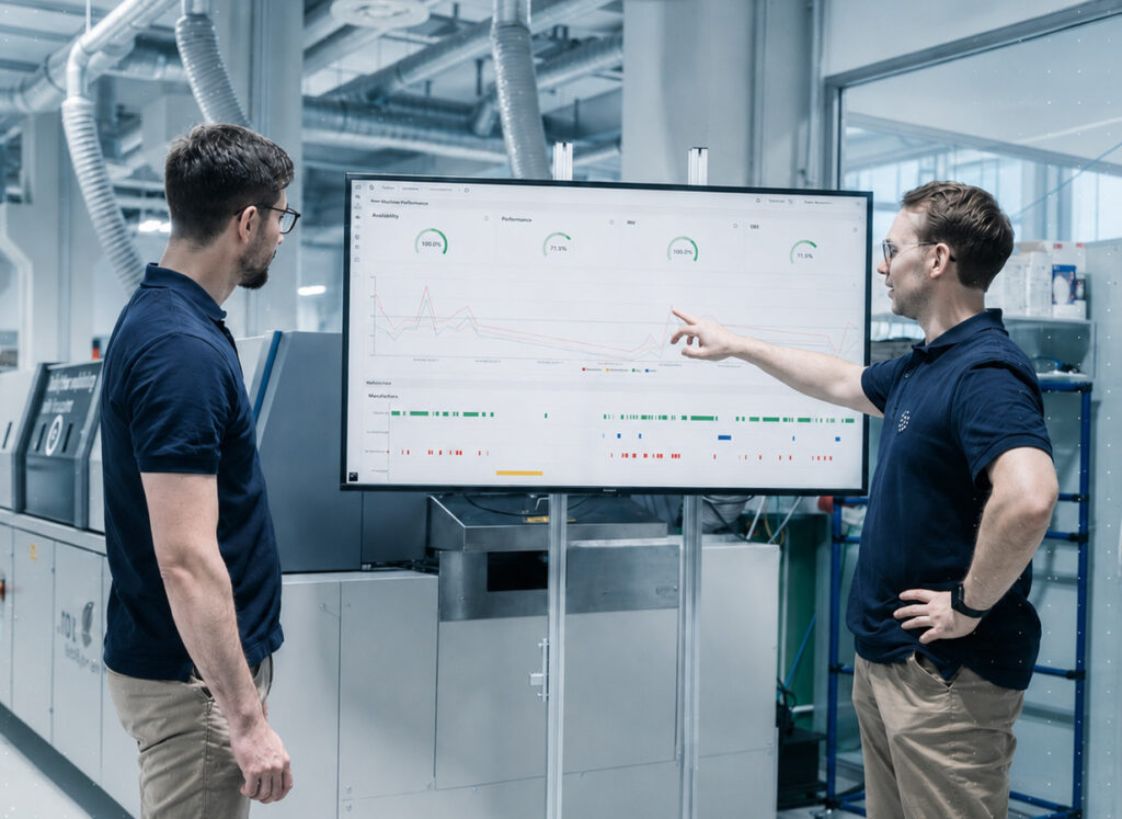 Zwei Techniker analysieren Daten auf einem Bildschirm in einer Fabrikhalle.